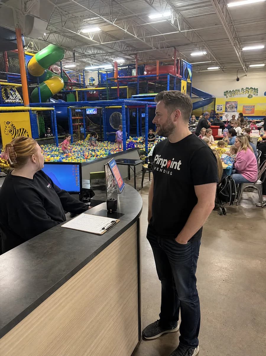 A Pinpoint rep in a branded tee standing at the front desk of an indoor playground, talking with the staff member behind the counter while families eat at tables beside the ball pit.