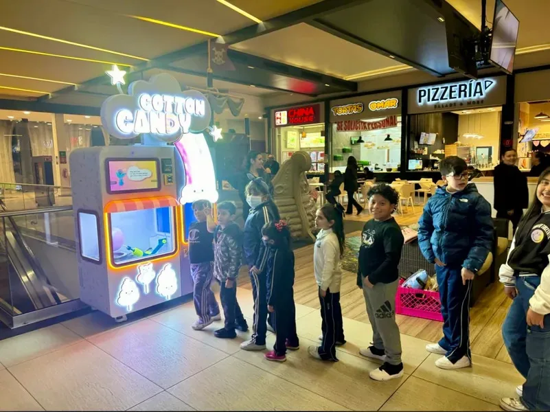 A lit cotton candy vending machine in a shopping mall food court, with a line of children waiting their turn and pizzeria and food counters behind.